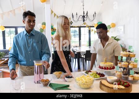Verschiedene Freunde treffen sich auf der Kücheninsel und wählen Cupcakes und rosa Drinks am Hausbuffet. Feier, Geselligkeit, Festlichkeit, Gourmet, Inneneinrichtung Stockfoto
