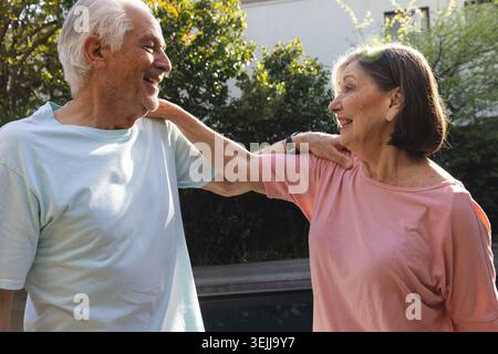 Seniorenpaar, das zusammen im Garten steht, lacht unter sonnendurchflutetem Laub mit Uhr. Ruhestand, Gesellschaft, Stockfoto