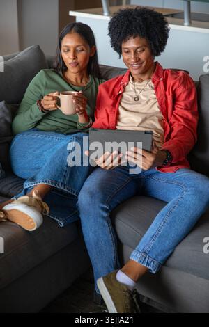 Indische und afroamerikanische Paare sitzen auf einem grauen Sofa im Wohnzimmer und halten ein Tablet und einen Becher. Modern, gemütlich, Lifestyle, mi Stockfoto