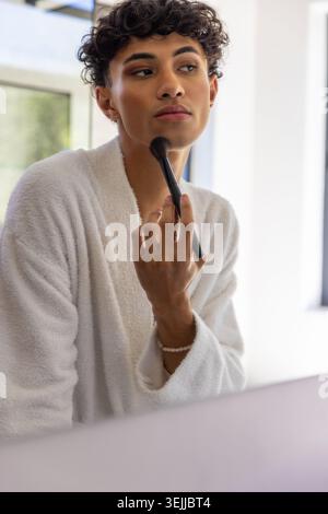 Hispanischer Mann in weißem Bademantel, der Make-up-Bürste auf das Kinn anwendet. Selbstpflege, Pflege, Hautpflege, W Stockfoto