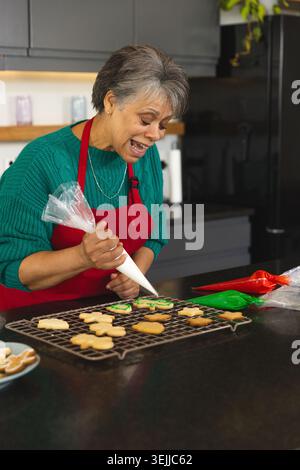 Ältere Frau in roter Schürze an der Küchentheke, die Plätzchen mit weißer Glasur dekoriert. Backen, Kuchendekorieren, Konf Stockfoto