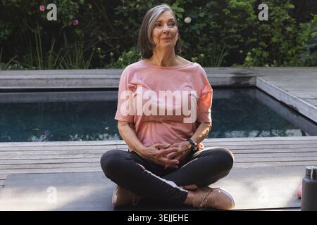 Seniorin in Sportbekleidung, die auf einer Matte an Deck sitzt und Yoga am Pool mit einer Wasserflasche praktiziert. Wellness, Entspannung, achtsam Stockfoto