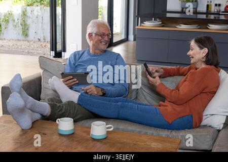 Seniorenpaar, das zu Hause auf einem grauen Sofa sitzt und ein Tablet und Smartphone in der Nähe von Kaffeetassen hält. Entspannung, Gesellschaft, modernes Int. Stockfoto