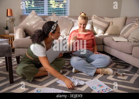 Verschiedene Freundinnen arbeiten mit gedruckten Karten und Laptop zusammen, die im Wohnzimmer auf Teppich sitzen. Zusammenarbeit, Teamwork, Crea Stockfoto