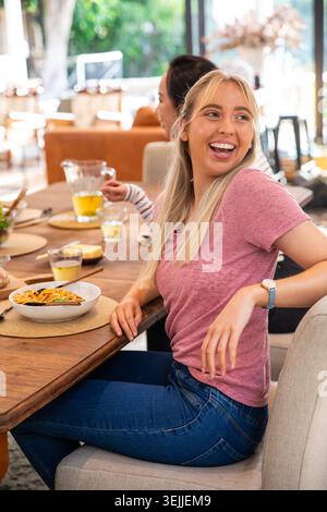 Verschiedene Freundinnen lachen und essen in einem hellen Essbereich mit Nudeln und Pitcher. Paare, Ladies, Lächeln, Pasta, dri Stockfoto