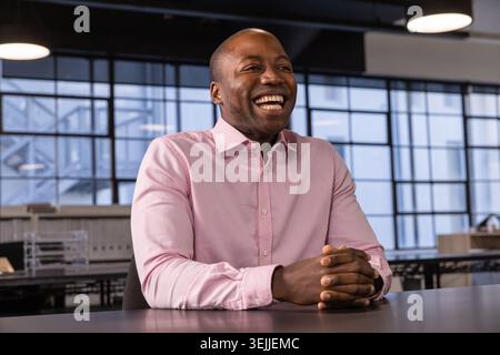 Lächelnder Afroamerikaner mit rosa Hemd, der sich vor dem Bürotisch mit Gitterfenstern beugt. Professionell, geschäftlich, Modus Stockfoto