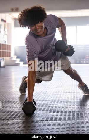 Afroamerikaner, der eine Planke im Fitnessstudio hält, während er eine Hexenhantel auf Gummiböden in der Nähe von Fenstern rudert. Fitness, Kraft, Gymnasium Stockfoto
