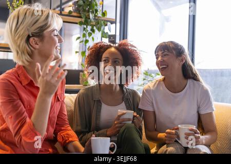 Verschiedene Freundinnen unterhalten sich, während sie in der Lounge auf einem gepolsterten Sofa sitzen und weiße Kaffeetassen halten. Lässig, Freundschaft, Entspannung Stockfoto