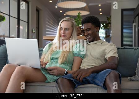 Verschiedene Paare mit silbernem Laptop auf grauem Sofa im modernen Wohnzimmer mit Hängelampen. Zusammenarbeit, Tragekomfort, Minim Stockfoto