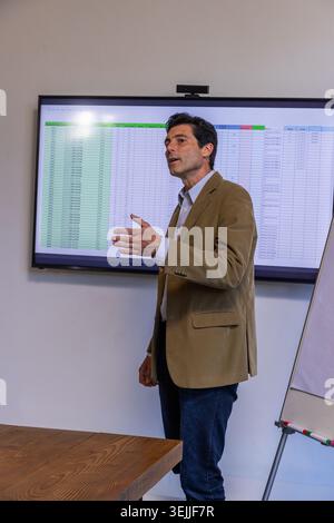 Mann mittleren Alters im Blazer, der Tabellenkalkulation auf einem Flachbildschirm im Büro mit Whiteboard präsentiert. Business, Präsentation, Profe Stockfoto