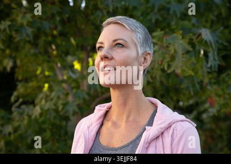 Frau, die zwischen Bäumen mit roten Beeren steht, trägt einen rosafarbenen Reißverschluss-Hoodie, ein graues Tanktop, das nach oben blickt. Abenteuer, Outdoor, Wellness Stockfoto
