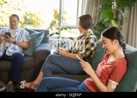 Vielfältige Familie, die Smartphones nutzt, während sie im Wohnzimmer mit Kissen und Fenster auf dem Sofa sitzt. Familienverbindung, Entspannung, moderner Lebensstil, gemütliche Umgebung Stockfoto