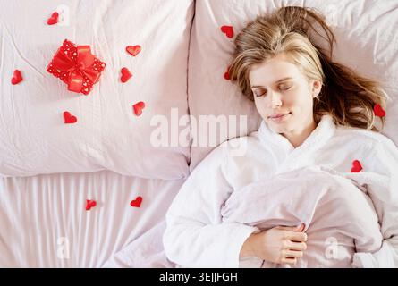 Junge blonde Frau im Bett mit Weihnachtsgeschenk und rotem Konfetti Stockfoto