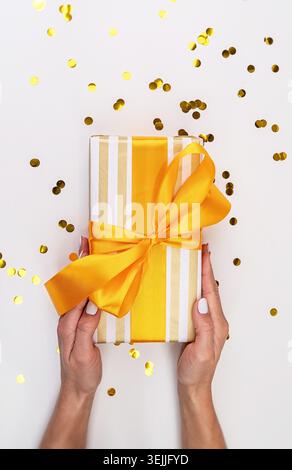 Eine Frau, die eine Geschenkbox in weiß-goldenem Papier mit verstreuten Konfetti hält Stockfoto
