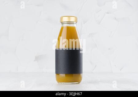 Orangefarbener Mangobaft in Glasflasche mit goldenem Verschluss und schwarzem leerem Etikett auf weißem Holztisch in hellem Innenraum mit Kopie s Stockfoto