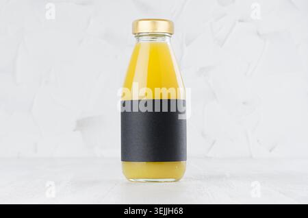 Orangefarbener, gelber Saft in Glasflasche mit goldenem Verschluss und schwarzem leerem Etikett auf weißem Holztisch in hellem Innenraum mit Kopie Stockfoto