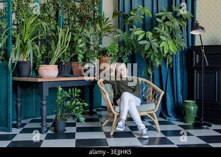 Junge blonde Frau, die in einem bequemen Stuhl sitzt, umgeben von Pflanzen Stockfoto