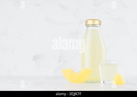 Gelber Bio-Ananassaft in Glasflasche mit Weinglas, Fruchtscheibe auf weißem Holztisch in hellem Innenraum, templ Stockfoto