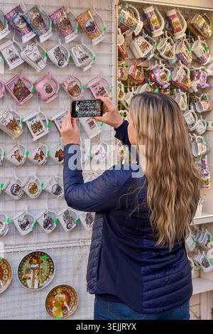 Eine blonde Frau fotografiert mit ihrem Smartphone eine mit bunten Kühlschrankmagneten bedeckte Erinnerungswand. Stockfoto