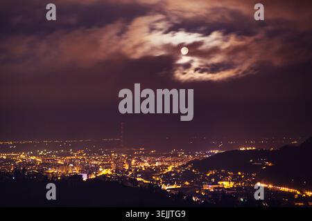 Leuchtende Stadtlichter beleuchten die Skyline der Stadt unter einem bewölkten Nachthimmel bei Vollmond Stockfoto