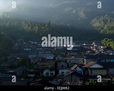 Blick aus der Vogelperspektive auf das Dorf Garung mit Menschen, die sich in der Nähe einer Moschee mit goldener Kuppel versammelt haben, während das Morgenlicht durch nebelgrüne Hügel in Kecamatan K fließt Stockfoto