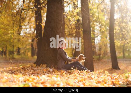 Junge schöne Frau, die im Herbst draußen mit Hund unter dem Baum sitzt Stockfoto