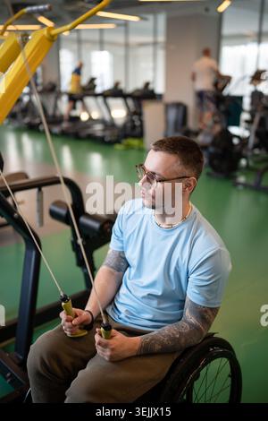 Entschlossener behinderter Mann in Rollstuhlzügen mit Fitnessgerät im Fitnessstudio, zeigt Ausdauer und Wunsch, in Zukunft wieder auf die Beine zu kommen Stockfoto
