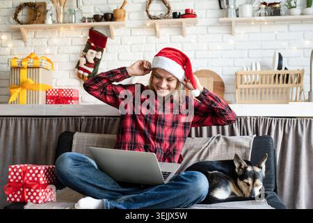 Glückliche junge Frau mit weihnachtsmann-Hut, die ihre Freunde im Video-Chat auf Laptop mit weihnachtsgeschenk begrüßt Stockfoto