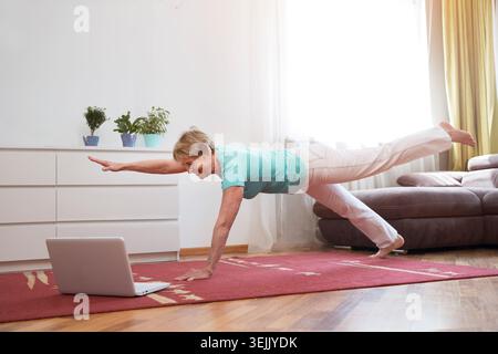 Aktive Seniorin, die Yoga-Fitnesstraining macht und Online-Workout-Tutorials auf dem Laptop im Wohnzimmer zu Hause ansieht. Startseite Stockfoto