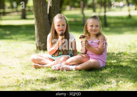 Zwei Schwestern sitzen auf Gras und essen Eis im Park an einem heißen Sommertag Stockfoto