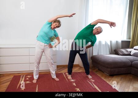 Aktives Seniorenpaar, das zu Hause Dehnübungen und Yoga macht. Fitness zu Hause, Aktivkleidung. Gesunder und sportlicher Lebensstil. Stockfoto
