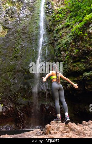 Frau, die auf Felsen steht, Arme offen, genießt einen malerischen Wasserfall in einer üppigen natürlichen Umgebung Stockfoto