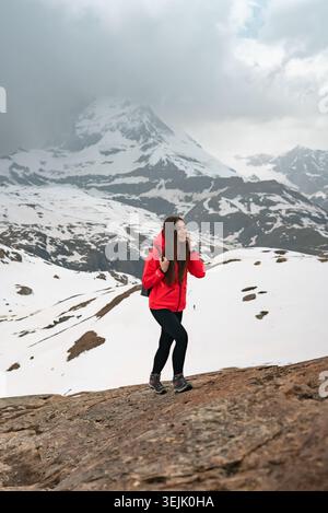 Frau in roter Jacke, die auf einem felsigen Weg mit verschneiten Bergen und matterhorn im Hintergrund wandert Stockfoto