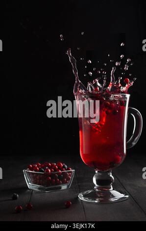 Rote Beeren Tee mit elegantem Spritzen und Wirbeln in Glas Weinglas mit reifer Cowberry, Heidelbeere auf schwarzem Holztisch in dunklem Blac Stockfoto