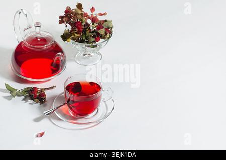 Herbsttee mit rotem Kräutertee in transparenter Teekanne, Tasse mit Weißdornbeeren, Hagebutte, trockene Blätter im Sonnenlicht auf weiß Stockfoto