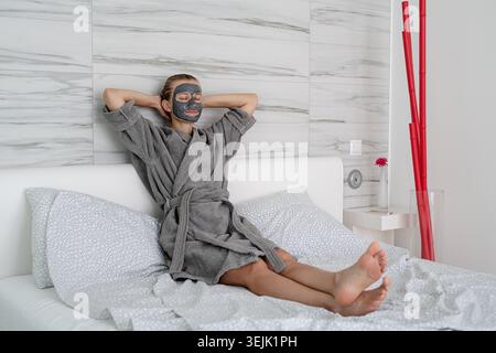 Frau mit Gesichtsmaske, die sich entspannt auf dem Bett sitzt Stockfoto