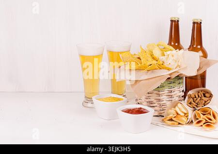 Sommer-Fast Food – verschiedene knusprige Snacks, zwei Lagerbier im Glas und braune Flaschen auf weicher weißer Holzplatte. Stockfoto