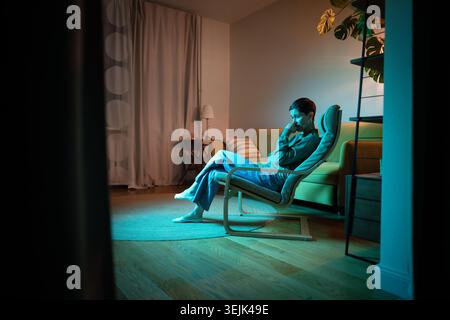Depressive, denkende Seniorin, die im dunklen Wohnzimmer auf einem Stuhl sitzt und sich in Denkprozessen einlässt. Stockfoto