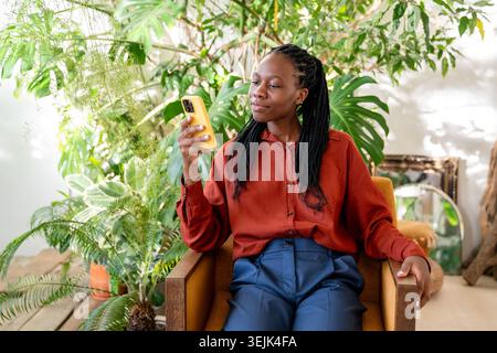 Positiv lächelnde afroamerikanische Frau, die sich im Stuhl mit Smartphone zu Hause im tropischen Garten entspannt. Stockfoto