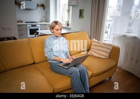 Entspannte Geschäftsfrau mittleren Alters, die auf einem Laptop tippt, während sie zu Hause auf einer bequemen Couch im Zimmer sitzt. Stockfoto