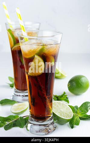 Kalter Cocktail cuba libre in zwei nassen langen Gläsern mit Strohhalm, Stücken Limette, Minze auf weichem, modernem weißem Hintergrund. Stockfoto