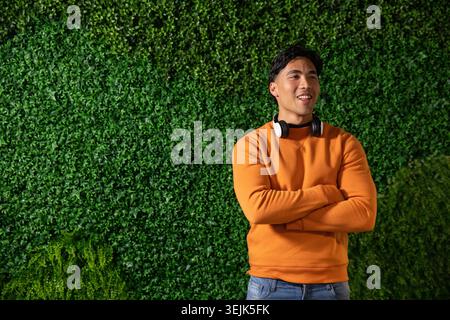 Asiatischer Mann, der an der grünen Wand steht, links in orangefarbenem Pullover und Kopfhörern blickt, Kopierraum. Laub, draußen, Porträt, mi Stockfoto