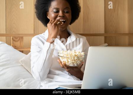 Faule schwarze Frau, die sich entspannt, Popcorn isst, während sie online Filme auf Laptop ansieht, im Bett liegend. Stockfoto