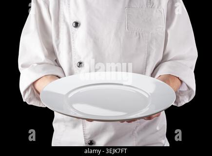 Chef in weißer Uniform hält einen leeren weißen runden Keramikservierteller. Schwarzer Hintergrund. Stockfoto