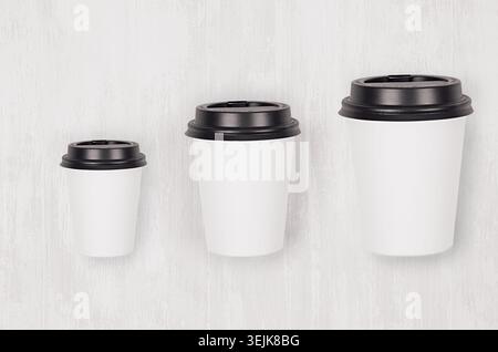 Coffee Mockup – Set aus drei verschiedenen Größen, weißen Papierbechern und leerer schwarzer Kappe auf weißem Holzbrett, Draufsicht. Modern elegant c Stockfoto