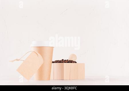 Kaffeepackung Mockup - Basteln Sie braune Pappbecher mit weißem Deckel, Kaffeebohnen im Mörtel und leeres Etikett auf weißem Holzbrett mit c Stockfoto