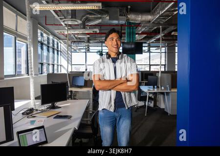 Asiatischer Mann, der im modernen Büro steht und lächelt, in gestreiftem Hemd mit Kopfhörern neben Monitoren. Arbeitsbereich, Tageslicht, Glas, Skyl Stockfoto