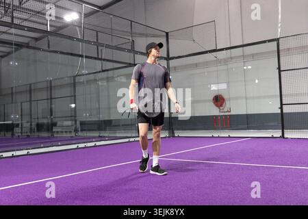 Männlicher Athlet, der auf dem violetten Padel-Court steht und einen perforierten Schläger hält und ein dunkelgraues Hemd trägt Stockfoto