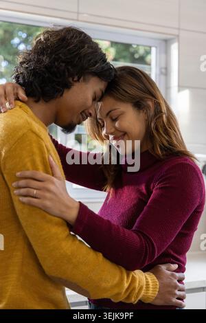 Ein Paar, das die Stirn am Küchenfenster drückt, Senf- und Burgunderstricke trägt, Ring auf der Brust. Intim, umarmen, sonnenbeleuchtet, Zuhause, Stockfoto