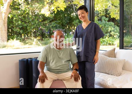 Afroamerikanische Seniorin sitzt auf rosa Ball zu Hause asiatische Krankenschwester in Peelings, die die Schulter stützen Stockfoto